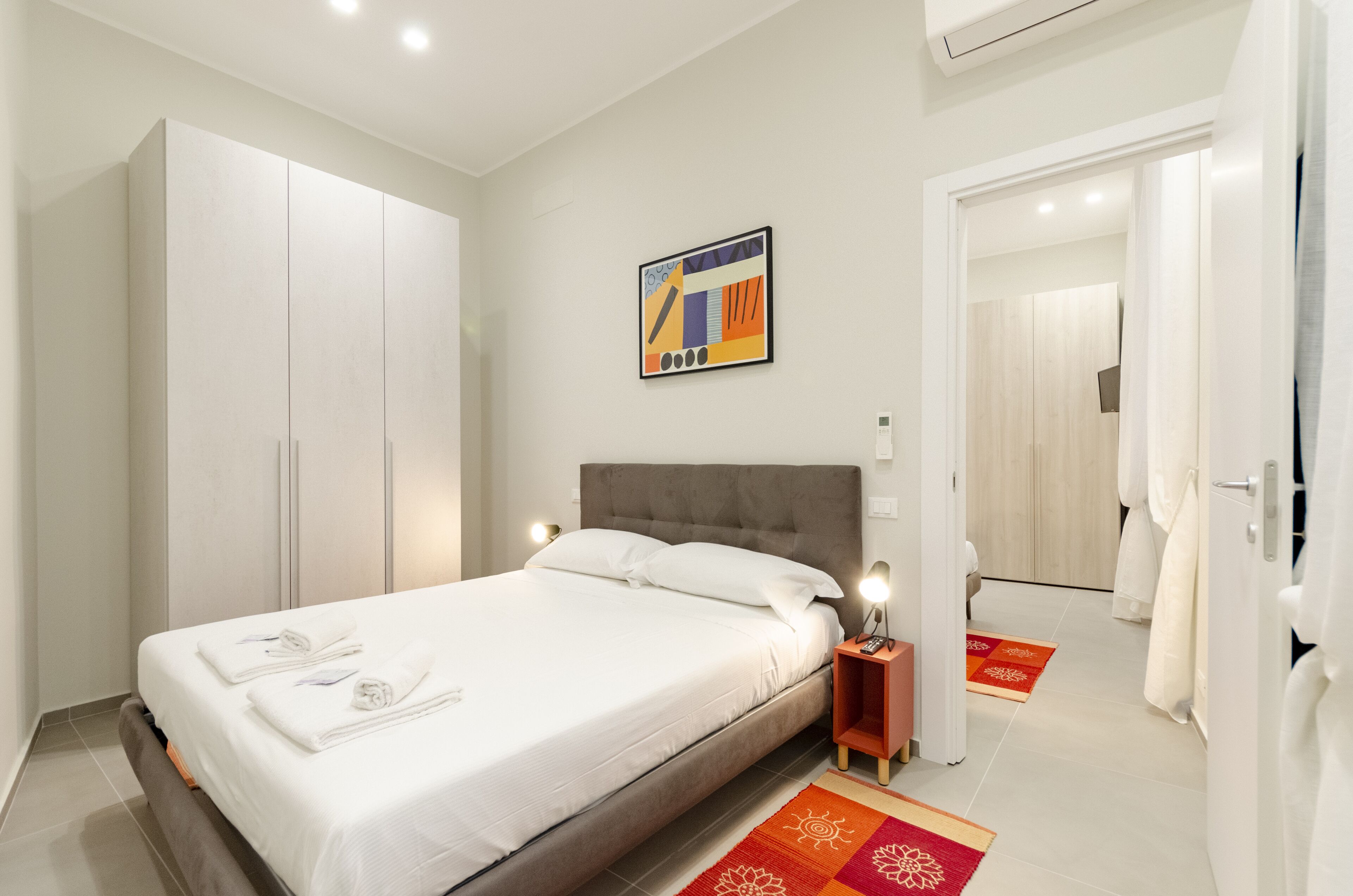Una habitación de hotel moderna con una cama grande, un armario, una mesita de noche con lámpara y una obra de arte colorida en la pared.