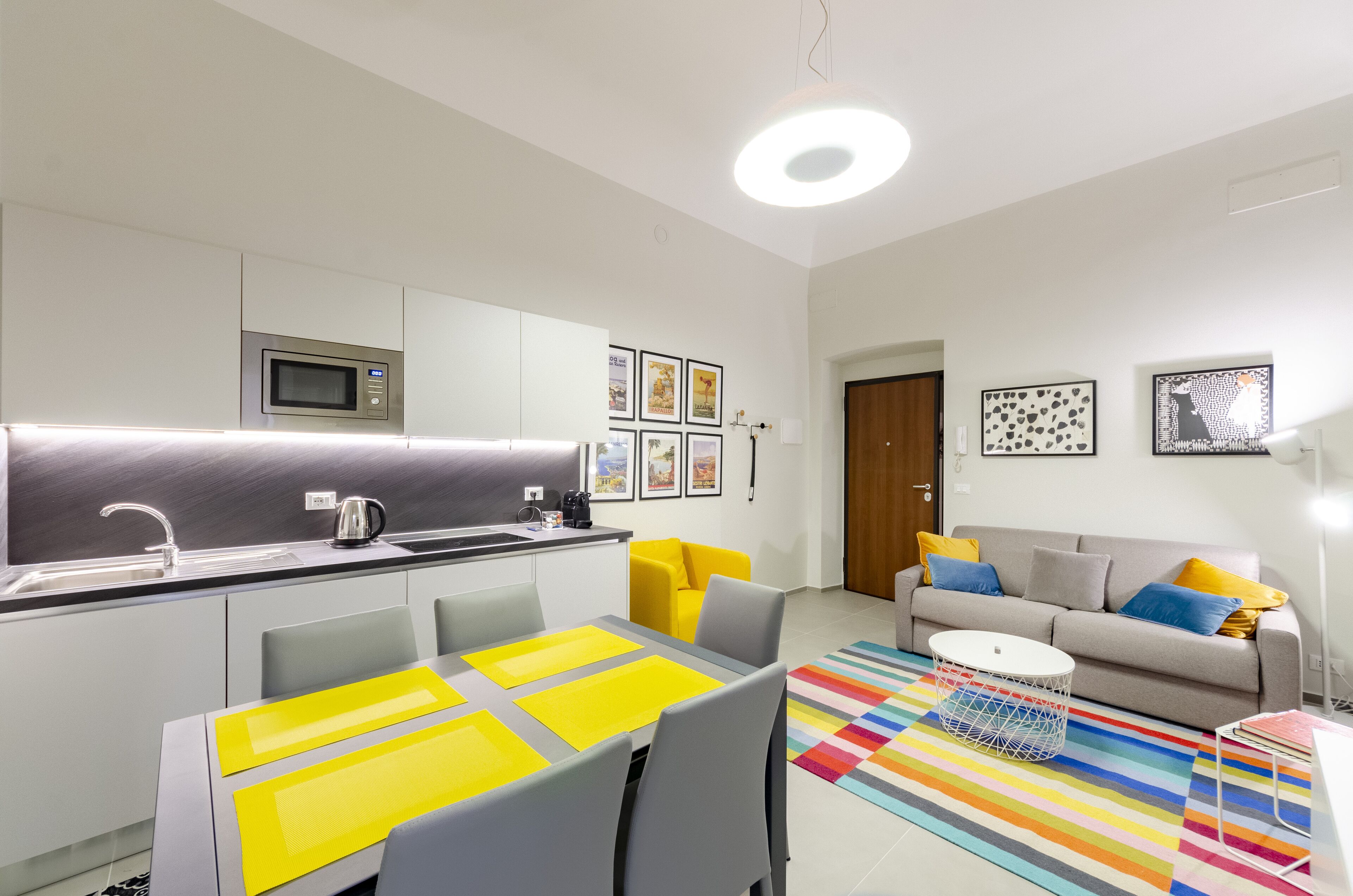 Une cuisine moderne équipée d’un four à micro-ondes, d’un évier et d’une bouilloire. On y trouve une table à manger avec des chaises et un tapis coloré. Un canapé et une lampe se trouvent dans l’espace salon.