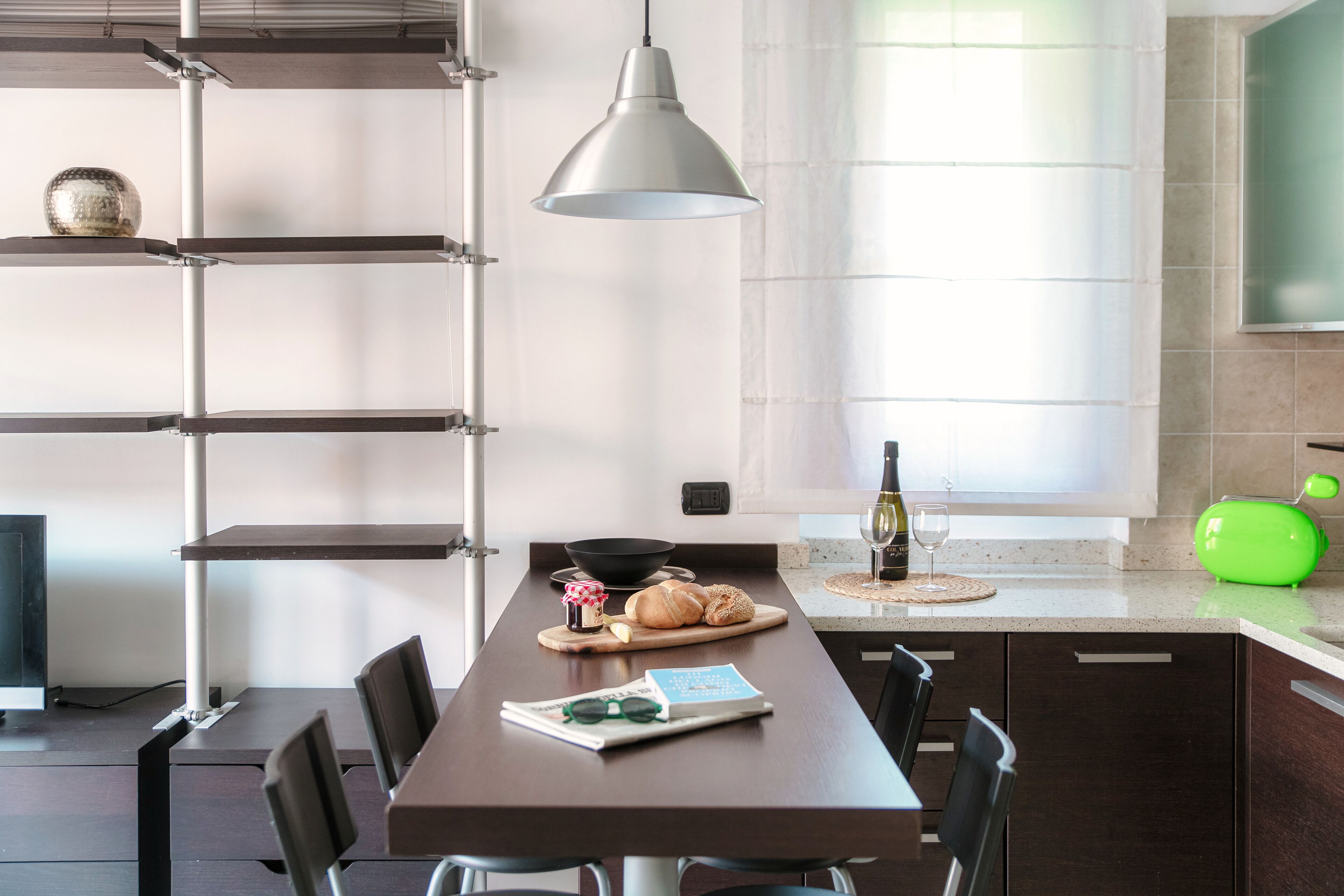 Una cocina moderna con una mesa larga, sillas, una lámpara colgante, un tostador verde y una copa de vino sobre la encimera.