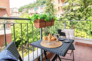 A balcony with a table set for breakfast, including croissants and a coffee pot, overlooking a residential area with greenery and buildings.