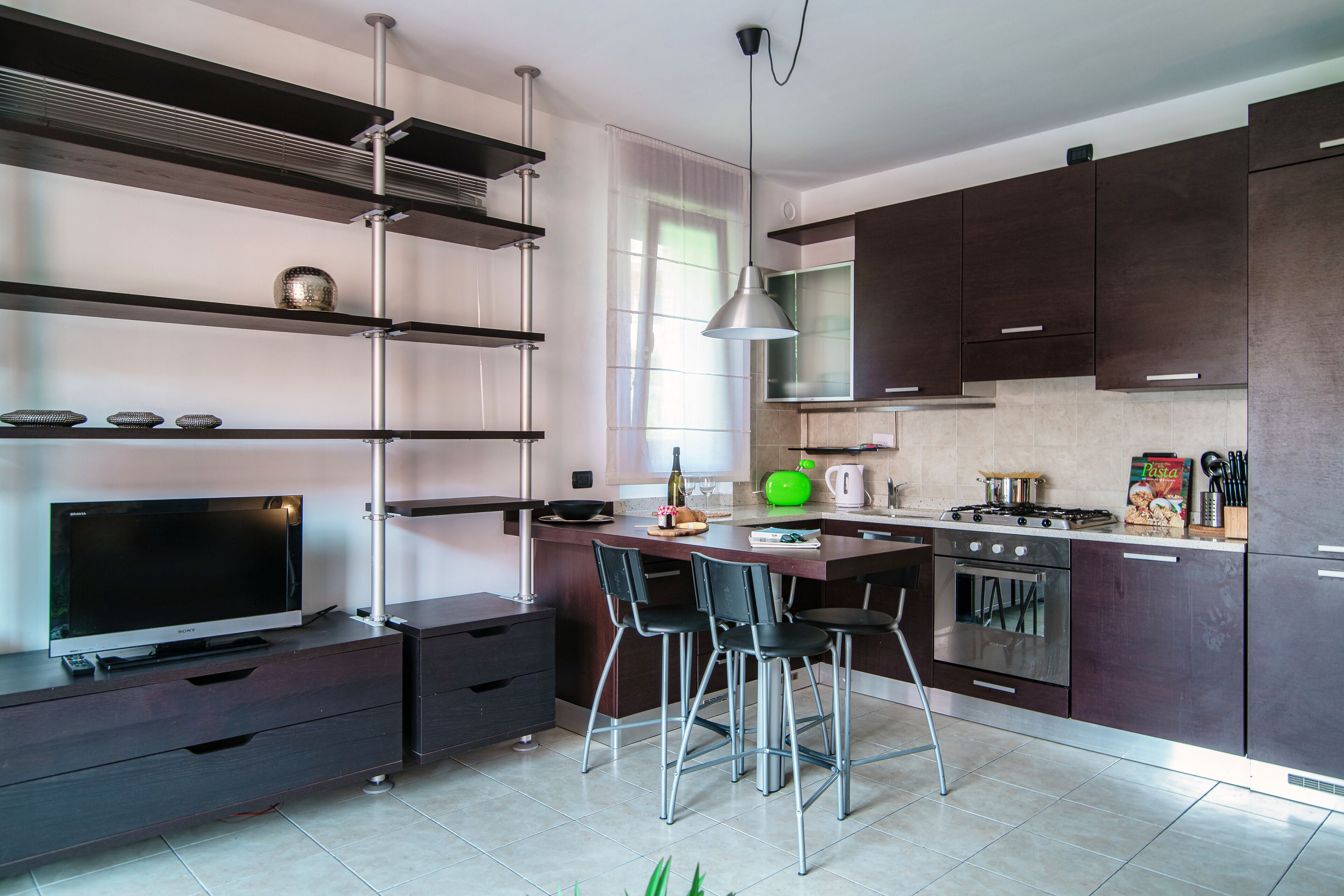 A modern kitchen with dark cabinets, a stainless steel range hood, a gas stove, a built-in oven, and a dining area with bar stools.