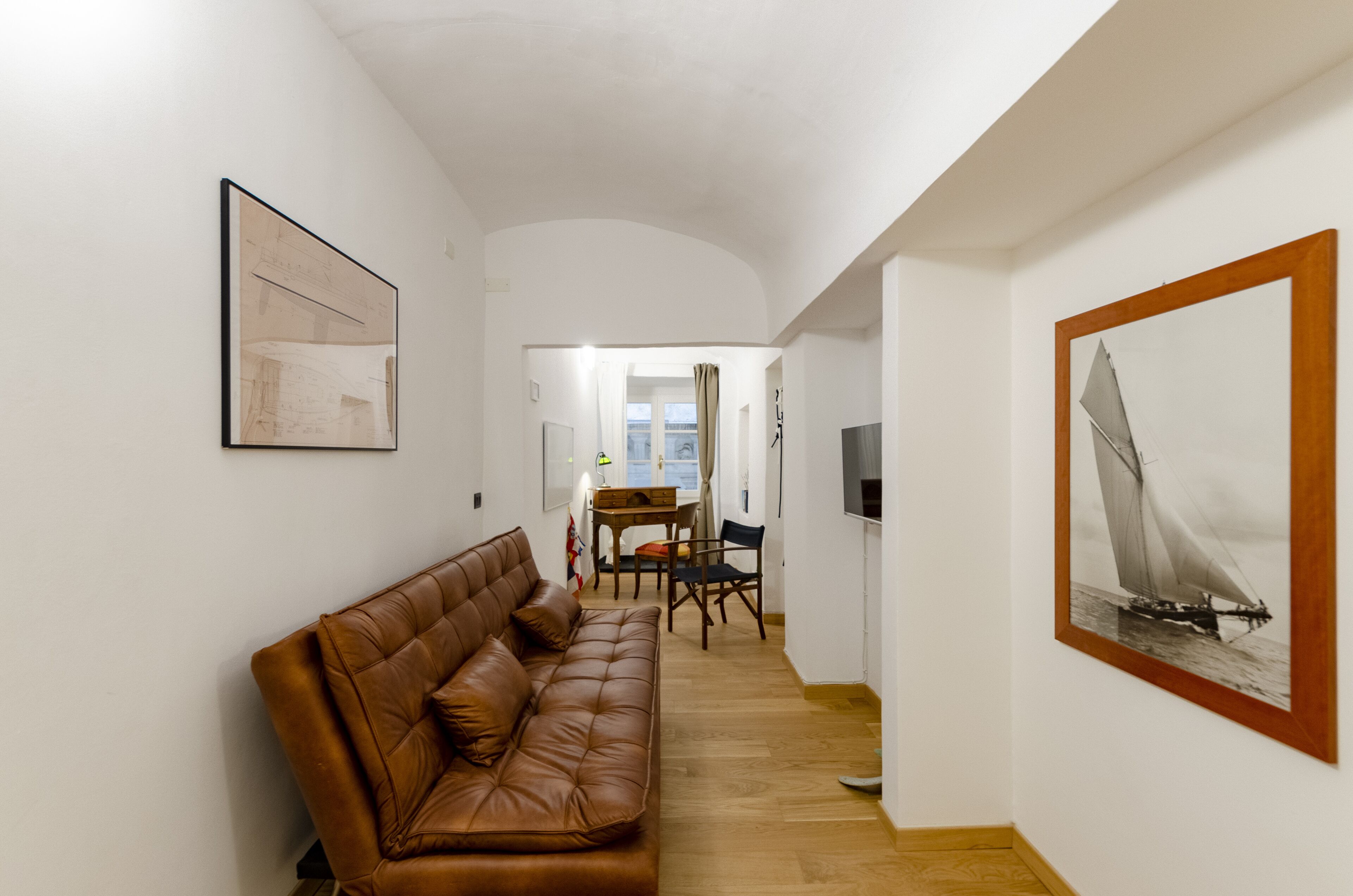 Un couloir avec un canapé en cuir marron, une table à manger avec des chaises, un téléviseur et un tableau encadré représentant un voilier.