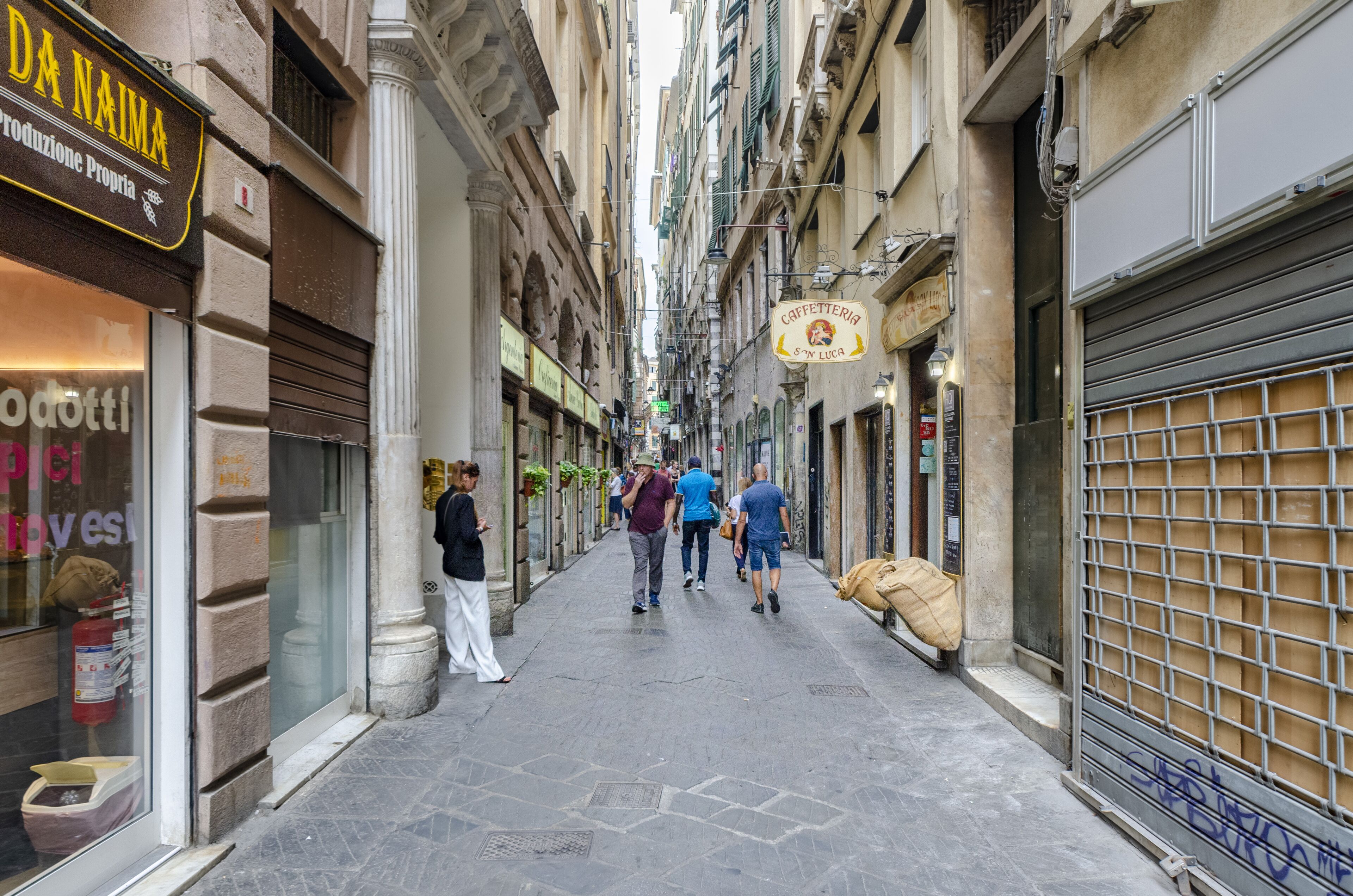 Una calle estrecha con tiendas y cafés, incluyendo un letrero visible que dice 'DA NAIMA' y 'Caffetteria S&M Luca'.