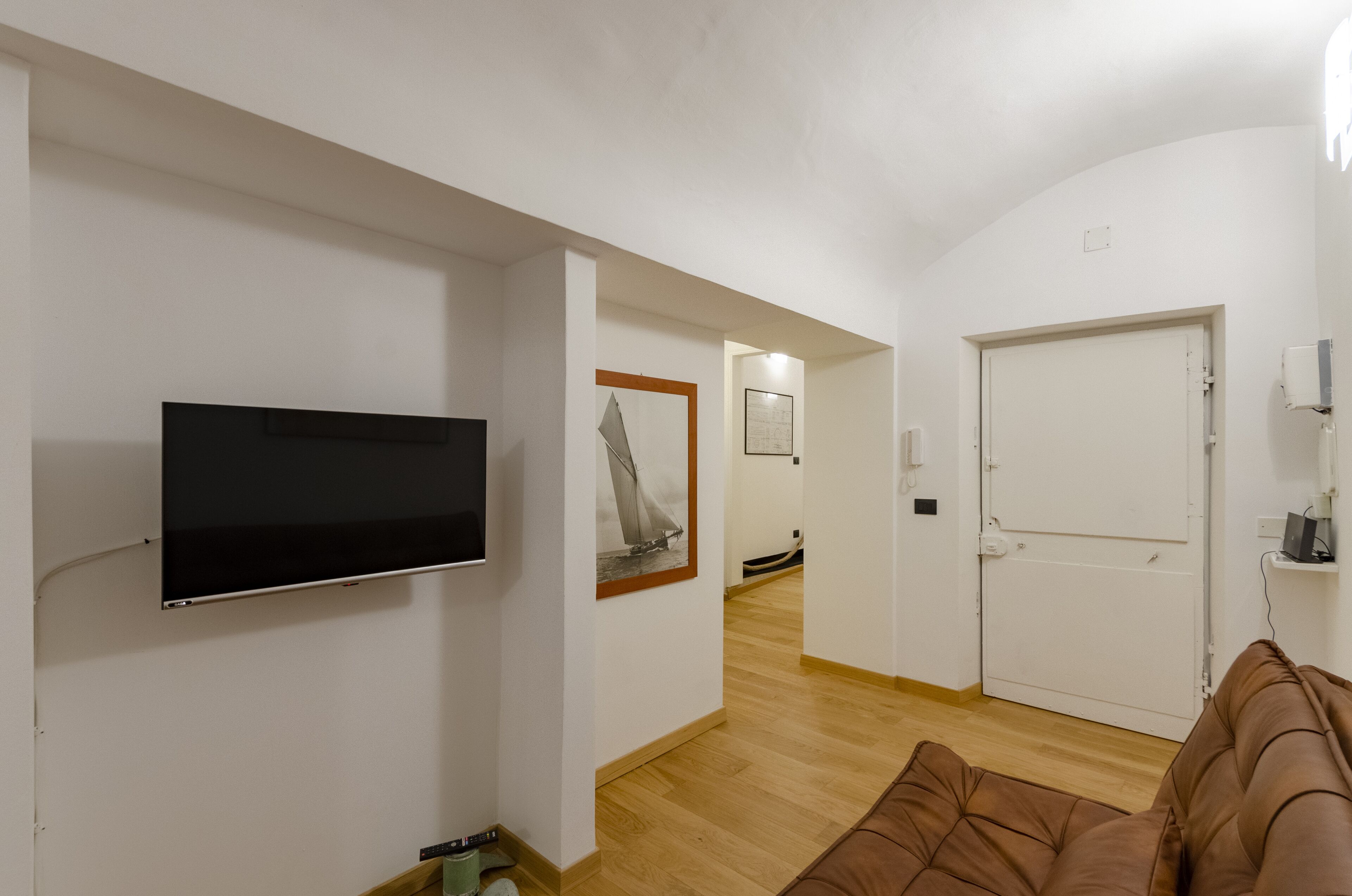 Ein Zimmer mit einem an der Wand montierten Flachbildfernseher, einer Tür, einem gerahmten Bild an der Wand und einem braunen Sofa.