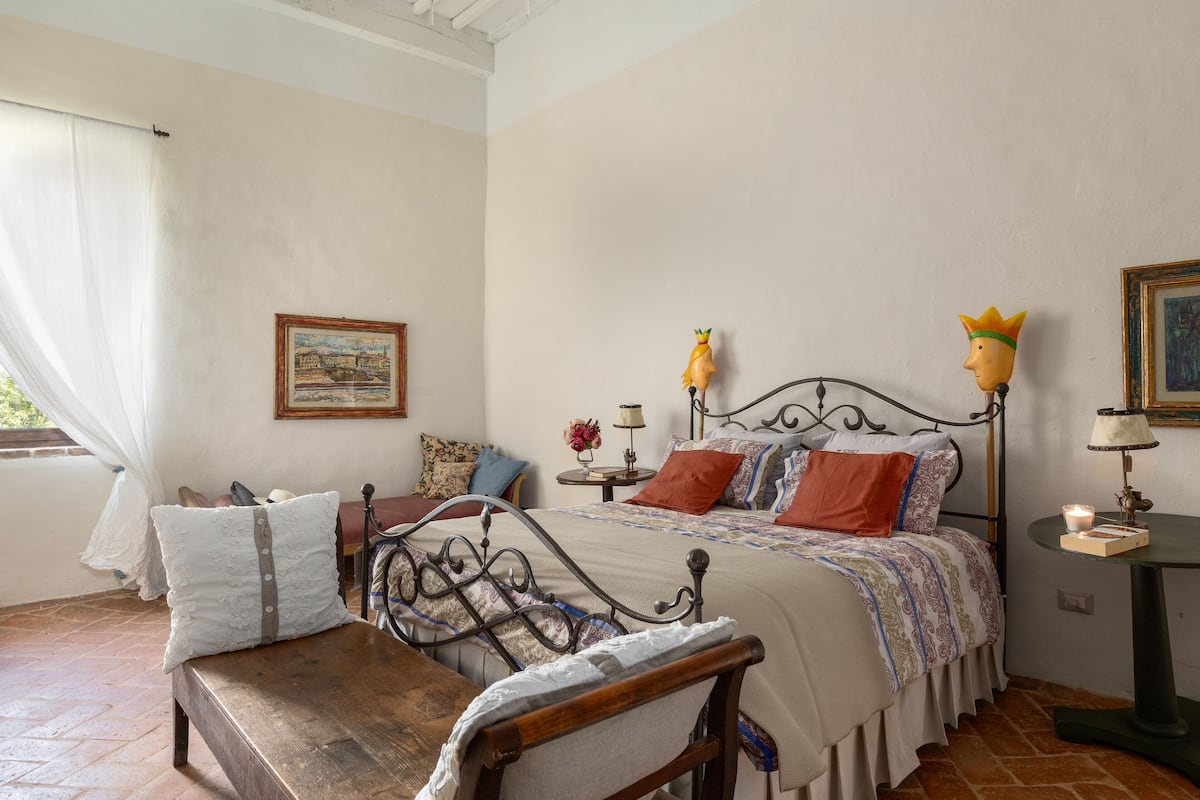 A bedroom with a bed, a wooden bench, a table, a lamp, and a framed picture on the wall.