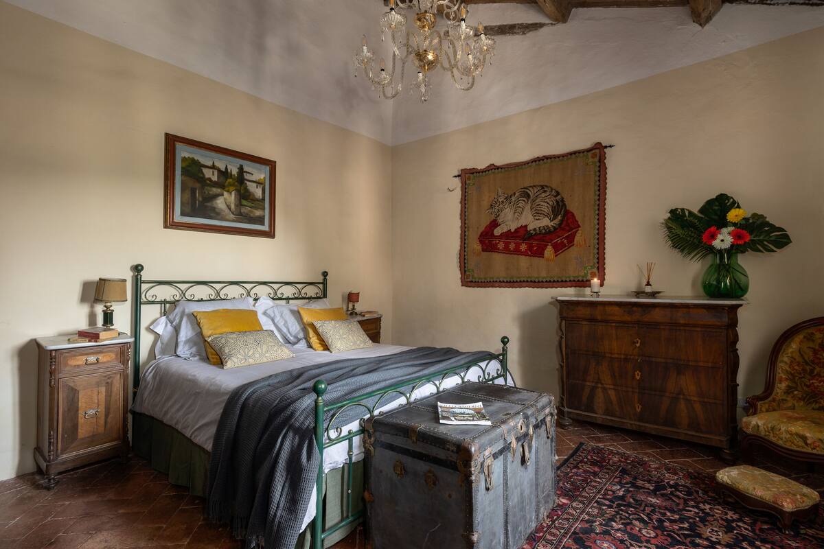 A bedroom with a bed, a dresser, a chair, a chandelier, and a painting on the wall.
