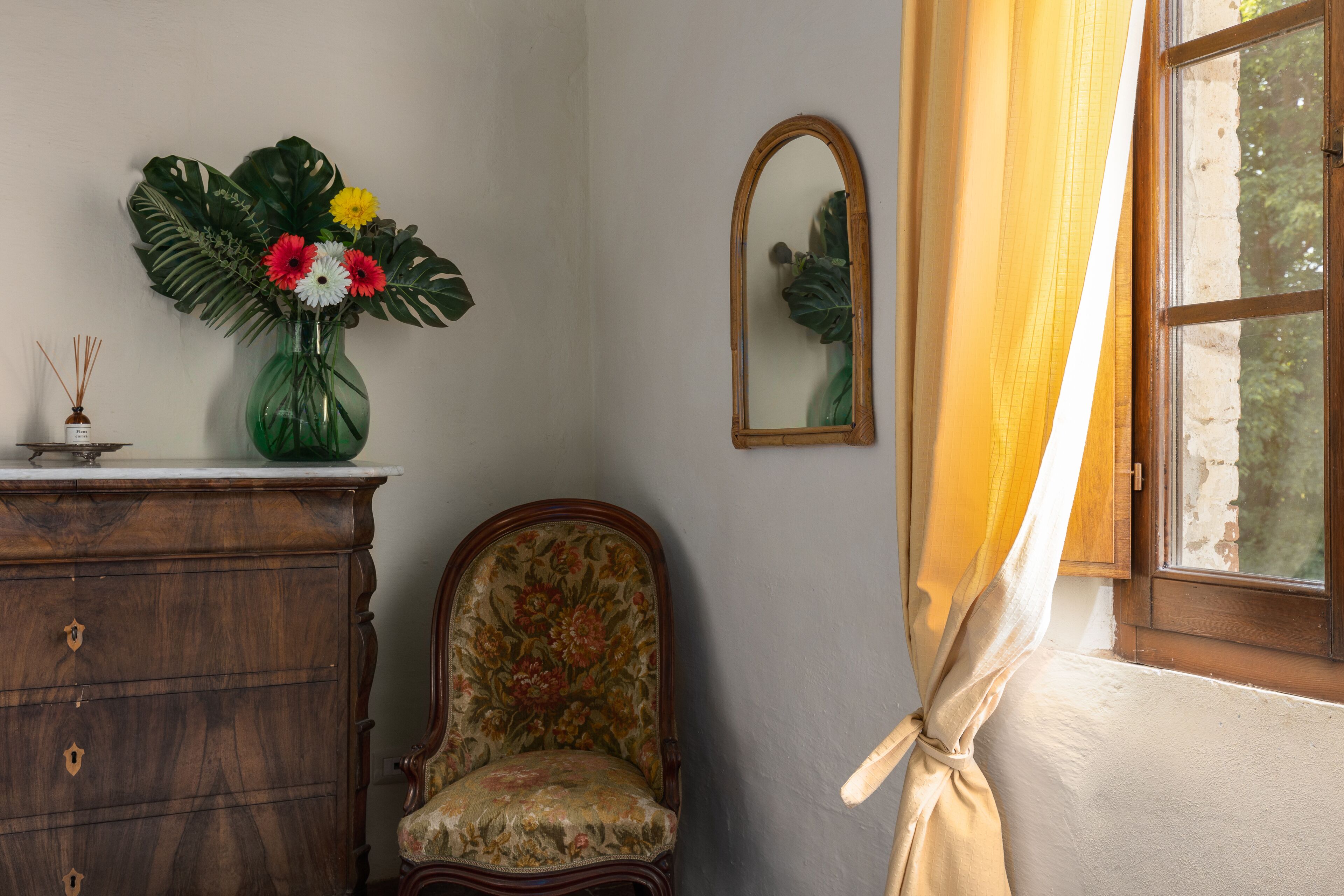 Une pièce comprenant une commode en bois, un fauteuil à motifs floraux, un vase vert avec des fleurs, un miroir et une fenêtre avec des rideaux.