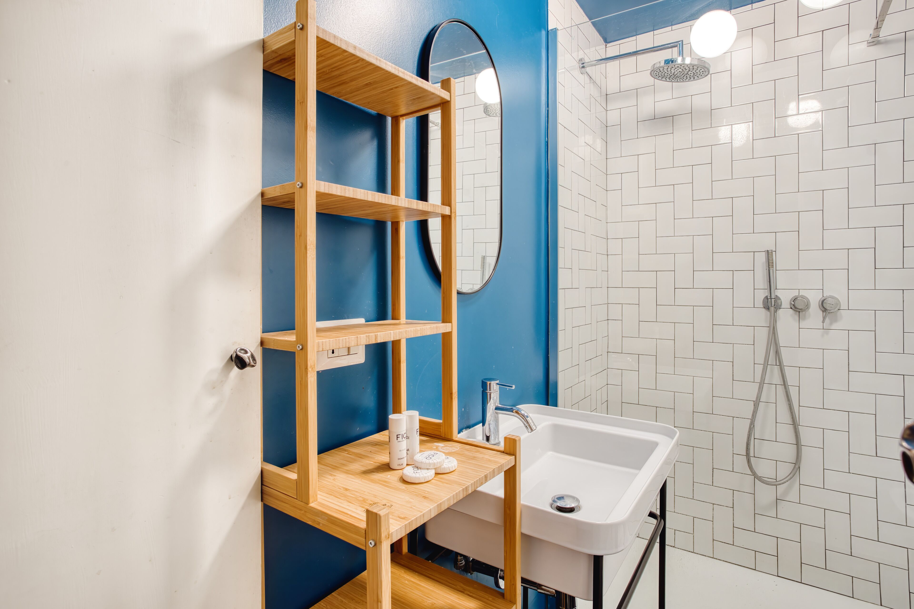 A bathroom with a white sink, a wooden shelf with toiletries, a mirror, and a shower area with white tiles.
