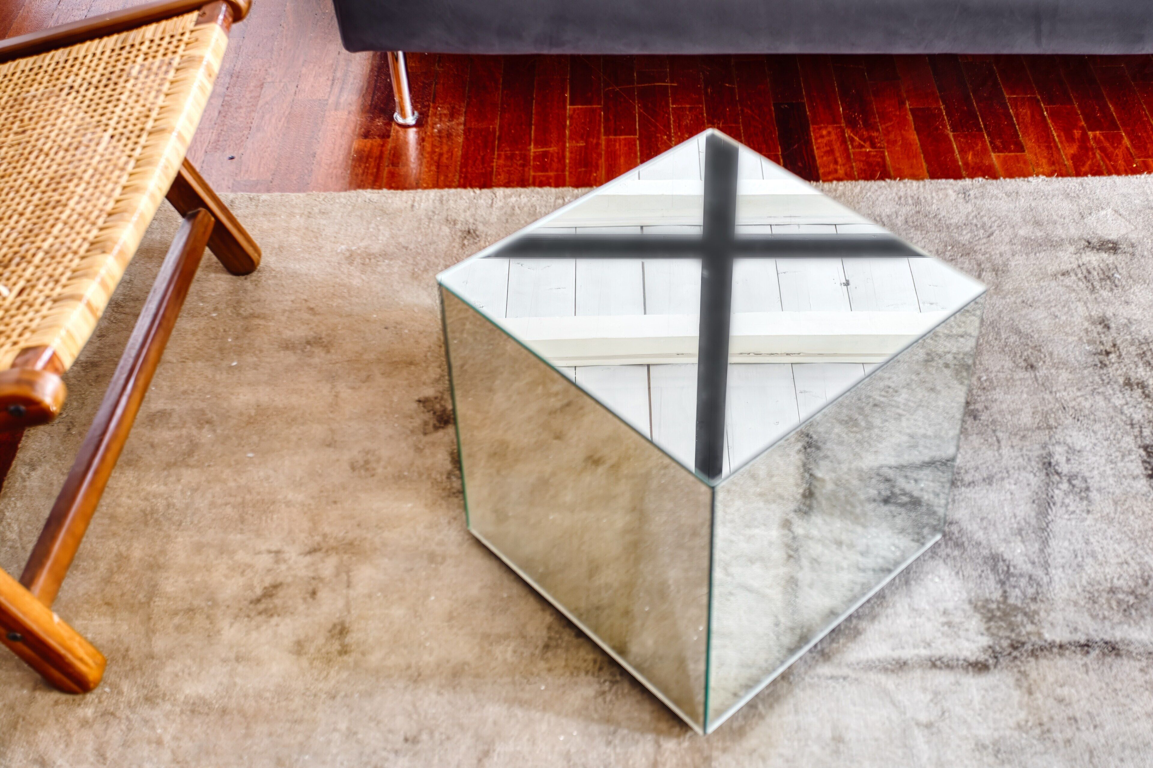 Une table basse en verre, ornée d’un motif en forme de croix, se trouve dans une pièce dont le sol est en bois et où est placée une chaise en osier.