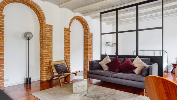 A modern living room with a grey sofa, wooden chair, and a glass coffee table. Brick arches and a large window are visible. - JOIVY Superb Studio In The Centre Of Milan (Milan)