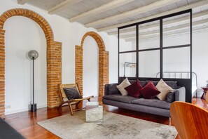 Un salon moderne avec un canapé gris, une chaise en bois et une table basse en verre. On peut voir des arches en briques et une grande fenêtre.