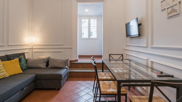A modern living room with a glass-top dining table, a grey sectional sofa, and a flat-screen TV mounted on the wall. - JOIVY Trevi Fountain's relais (Rome)