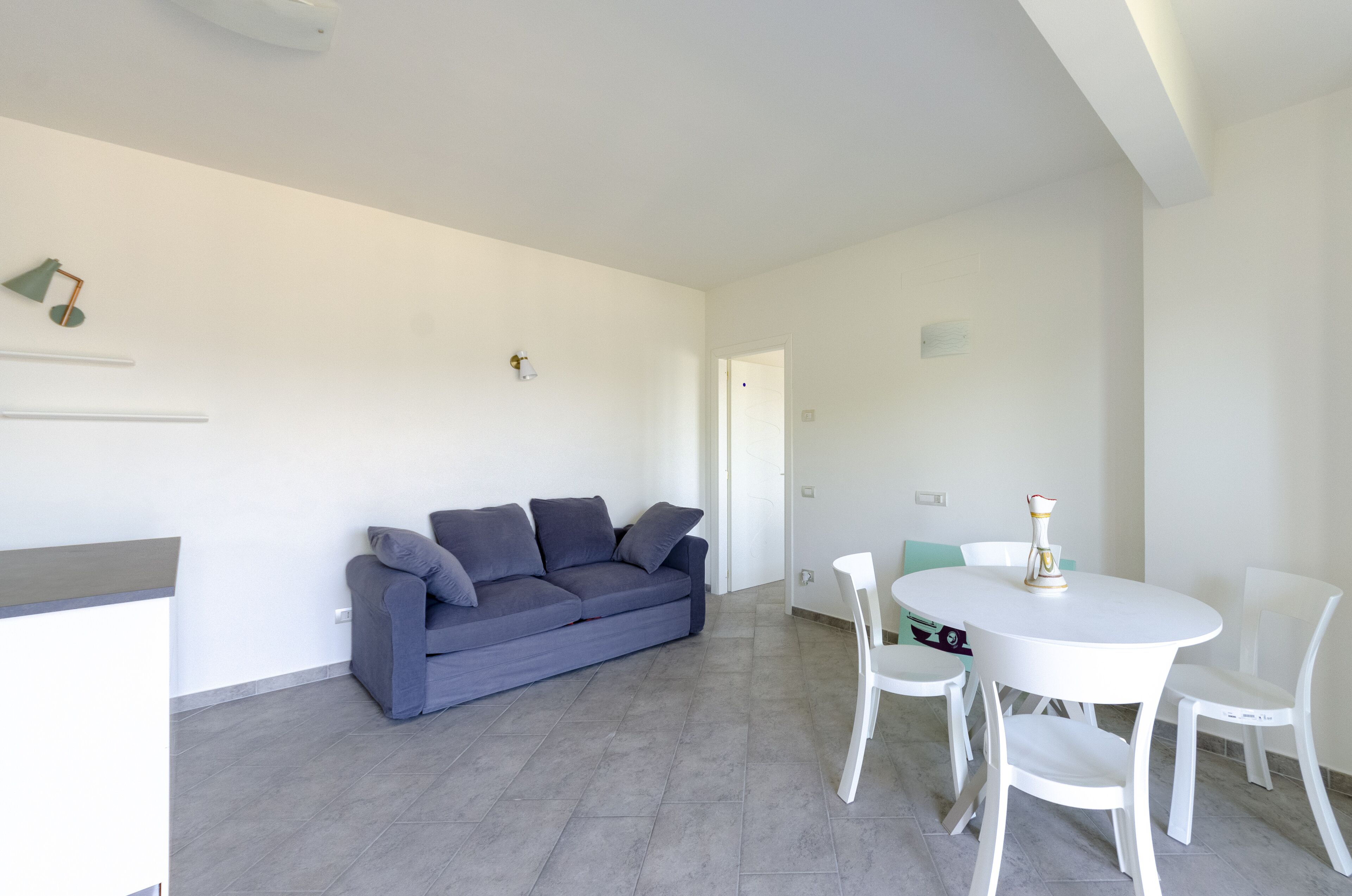 Una sala de estar moderna con un sofá gris, una mesa de comedor blanca con sillas y una lámpara decorativa pequeña.