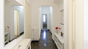 A narrow hallway with a white sink, mirror, and radiator. There are flower arrangements on the wall and a door at the end leading to another room.