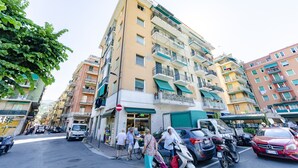 A multi-story residential building with balconies, a ground-level shop, and parked cars.