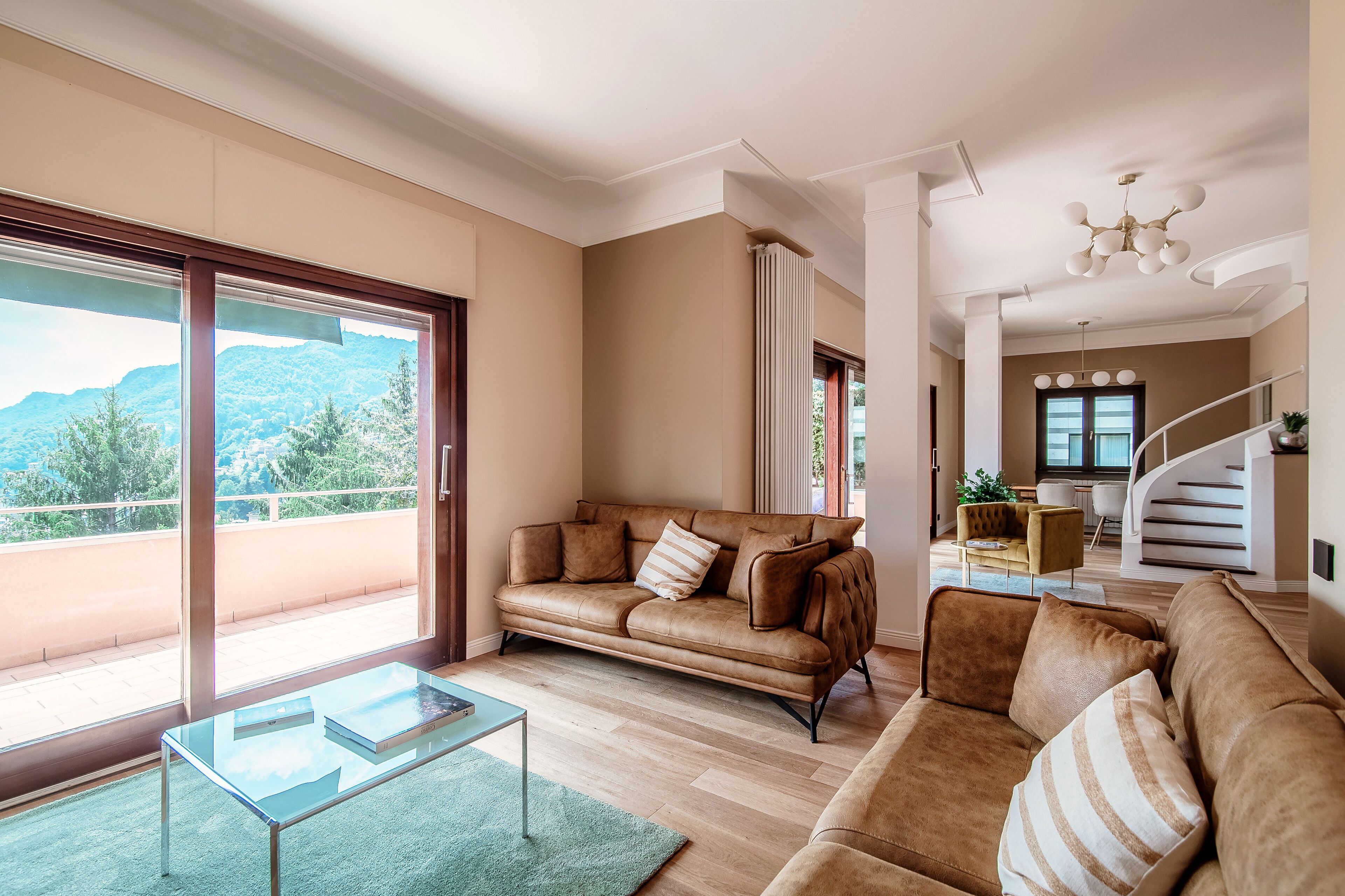 A spacious living room with a leather sofa, a glass coffee table, and a view of mountains through large sliding doors.