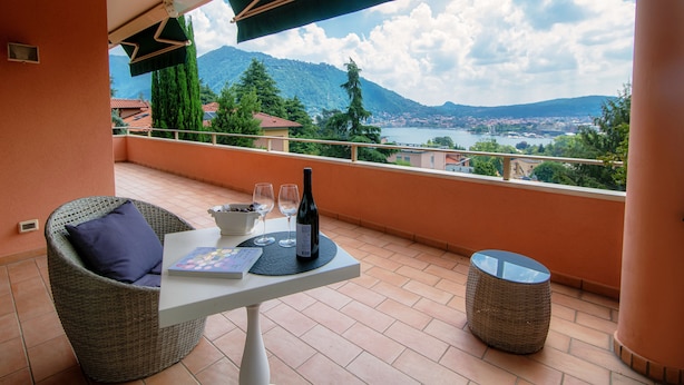 Un balcón con una silla de mimbre, una mesa con una botella de vino, dos copas y un libro. El balcón ofrece vistas panorámicas de montañas y un lago.
