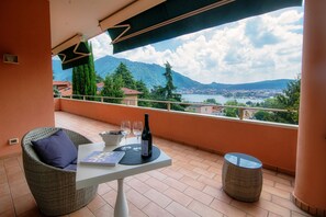 Ein Balkon mit Korbstuhl, Tisch, einer Weinflasche, zwei Gläsern und einem Buch. Der Balkon bietet einen schönen Blick auf Berge und einen See.
