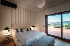A bedroom with a large bed, two bedside tables, a modern light fixture, and a view of a city through large windows.