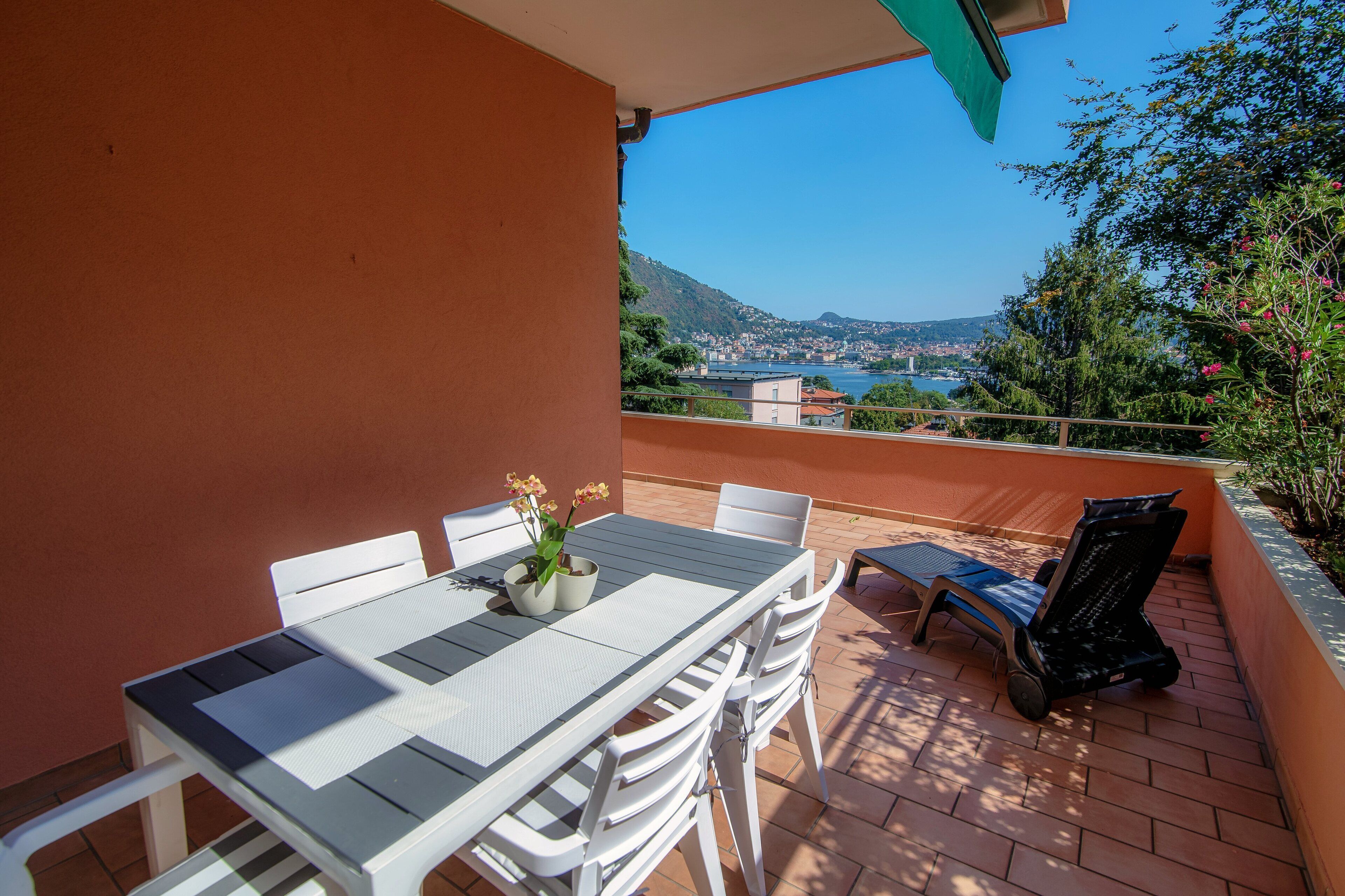 A terrace with a table and chairs, a potted plant, and a view of a body of water and hills.