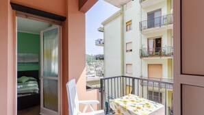 A balcony with a table and chairs, a bed with a green headboard, and a view of neighboring buildings.