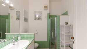 A bathroom with a green-tiled shower, a white sink, and a toilet. There are shelves with toiletries and a mirror above the sink.