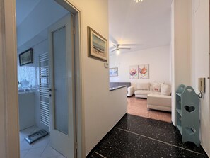 A hallway with a white door leading to a bathroom, a white sofa in a living area, and a ceiling fan. - JOIVY Blue Liguria (Rapallo)