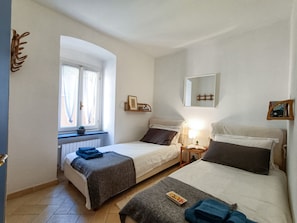A bedroom with two beds, a window, a bedside table with a lamp, and a mirror on the wall.