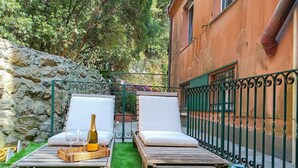 Zwei Liegestühle mit Kissen, eine Champagnerflasche und eine Snackplatte auf einem Holztisch in einem Garten mit grüner Wiese und einer Steinmauer.