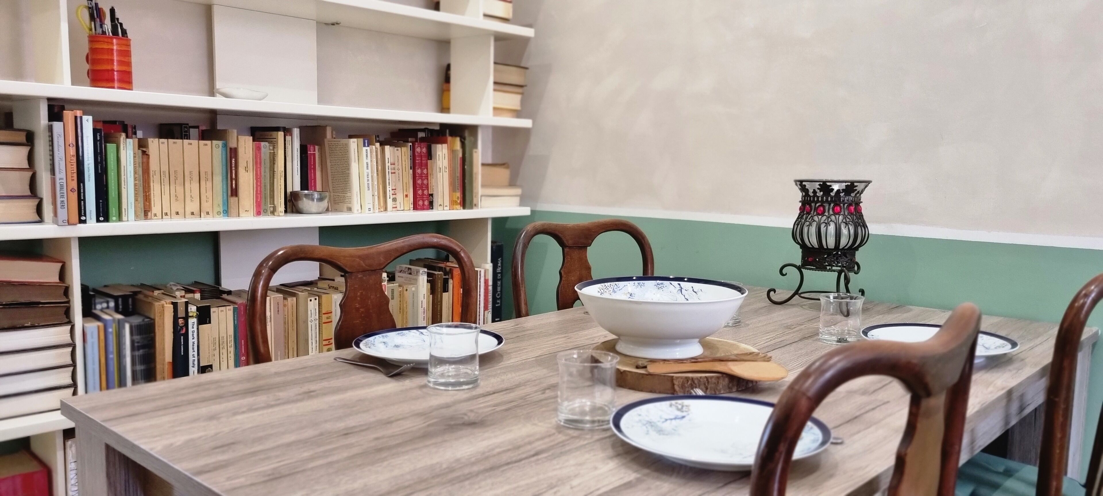 Une table à manger avec des chaises, un saladier, des assiettes et des verres. On aperçoit, à l’arrière-plan, des étagères remplies de livres et un vase décoratif.