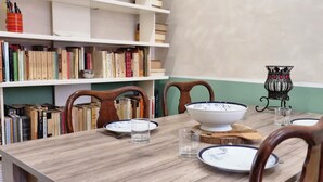 Ein Esstisch mit Stühlen, einer Schüssel, Tellern und Gläsern. Im Hintergrund sind Bücherregale mit Büchern und eine Ziervase zu sehen.