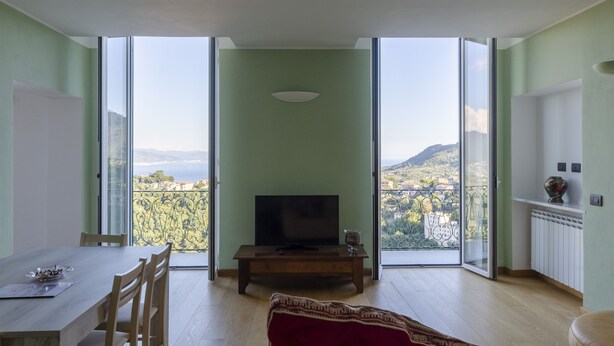Ein Wohnzimmer mit Fernseher, einem Esstisch und Stühlen. Ein großes Fenster bietet Blick auf einen Berg und einen See.