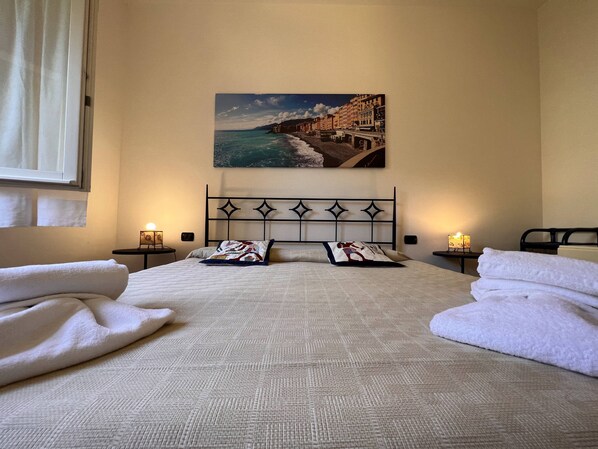 A neatly made bed with a patterned bedspread and pillows, two bedside tables with lamps, a painting of a coastal town on the wall, and a window with curtains.