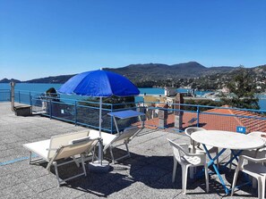 A terrace with outdoor seating, a blue umbrella, and a view of a lake and town in the background.