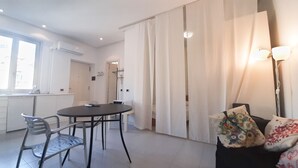 Ein modernes Wohnzimmer mit einem runden Tisch und zwei Stühlen, einem Sofa mit gemusterten Kissen und einem großen Fenster mit Vorhängen.