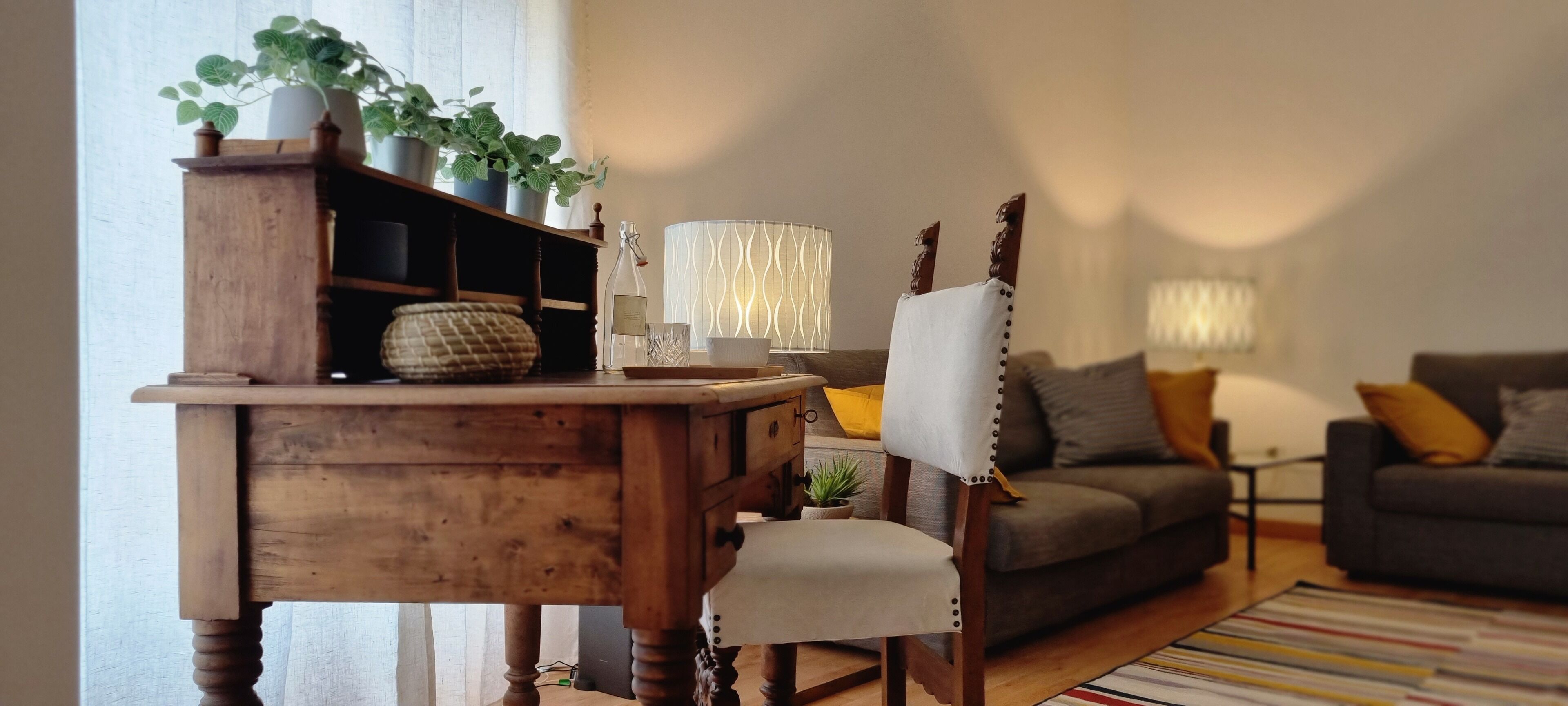 A wooden desk with a chair, a lamp, and a potted plant. There is a sofa and a rug in the room.