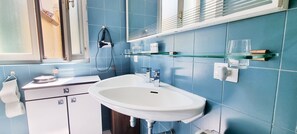 Un bagno con un lavandino bianco, piastrelle blu alle pareti, uno specchio e una finestra con persiane.