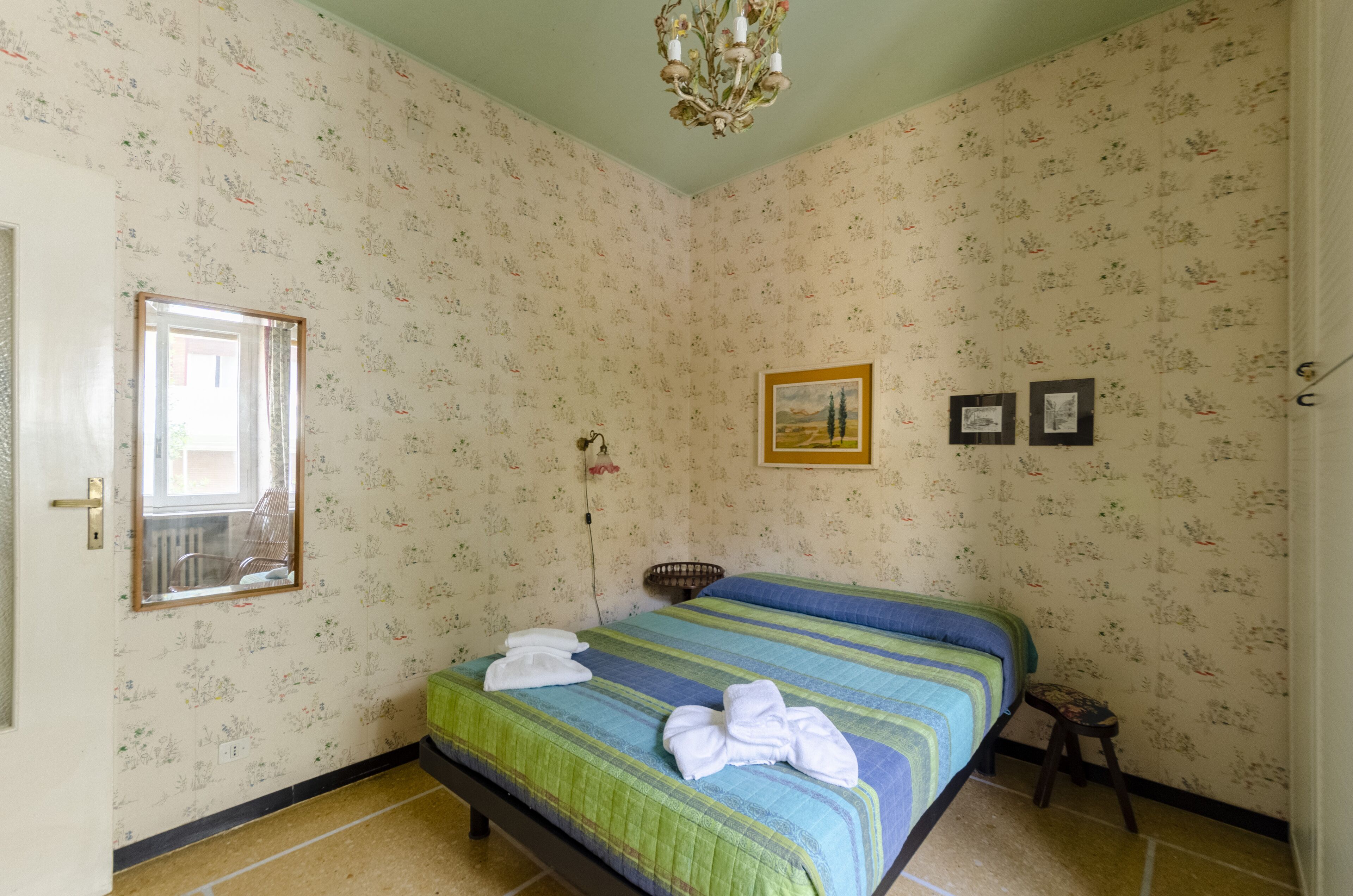 Une pièce avec un papier peint à motifs floraux, un lit avec une literie à rayures, un petit tabouret et un tableau encadré accroché au mur.