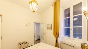 Ein Hotelzimmer mit einem Bett, einem Nachttisch, einem Fenster mit Vorhängen, einer Kristallleuchtre und einer Tür zu einem anderen Raum.