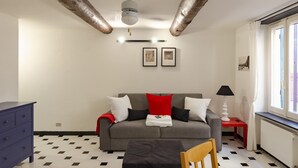 A room with a grey sofa, a red and white blanket, a wooden chair, a blue dresser, and a window with curtains. - JOIVY Flat By The Sea Promenade (Camogli)
