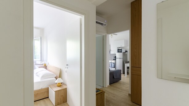 A modern hotel room with a bed, bedside table, and a small wooden nightstand with a lamp. A wooden wardrobe is visible to the right.