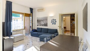 Ett vardagsrum med en soffa, ett matbord och en tv. Det finns ett fönster med gardiner, en väggmonterad luftkonditioneringsanläggning och en bokhylla.