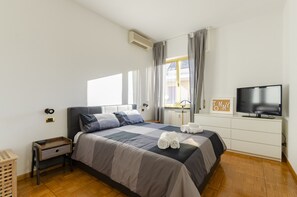 A neatly arranged bedroom with a bed, a TV, a bedside table, a lamp, and a window with curtains. - JOIVY Central Liberty Blue (Rapallo)
