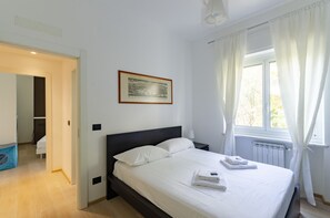 Ein ordentlich bezogenes Bett mit weißen Leinen, ein gerahmtes Bild an der Wand und ein Fenster mit Vorhängen.