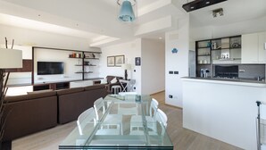 A modern living room with a glass dining table, brown sofa, and a kitchen area with a built-in stove. - JOIVY Great Flat W/Sea View (Genoa)