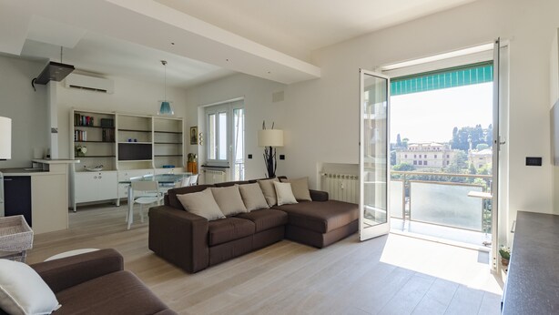 A modern living room with a sectional sofa, a dining area, and a kitchen. There is a large sliding glass door leading to a balcony with a view of buildings. - JOIVY Great Flat W/Sea View (Genoa)