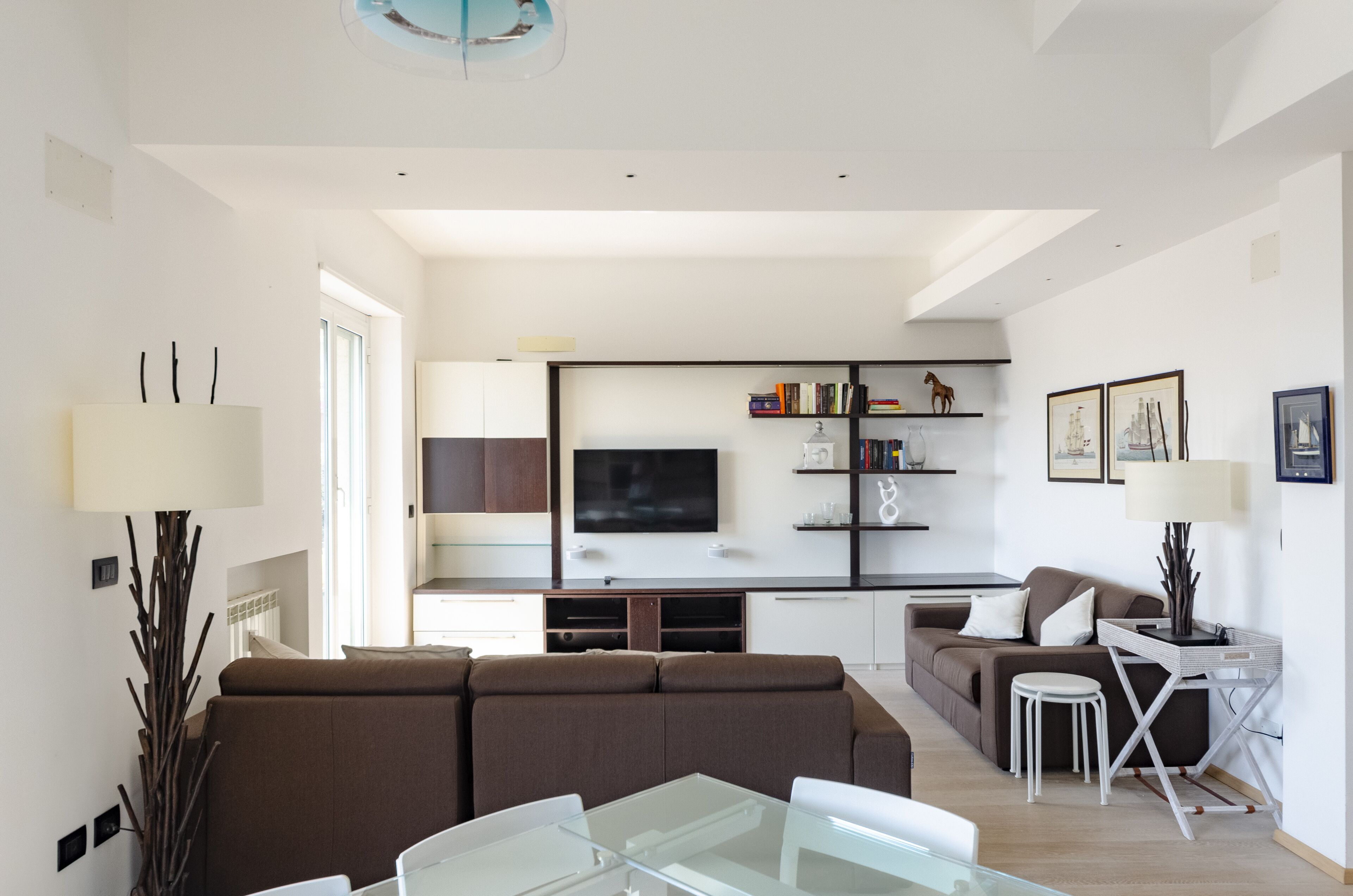 A modern living room with a sofa, a glass coffee table, and a TV. There is a kitchen area with a TV, bookshelves, and a dining table with chairs.