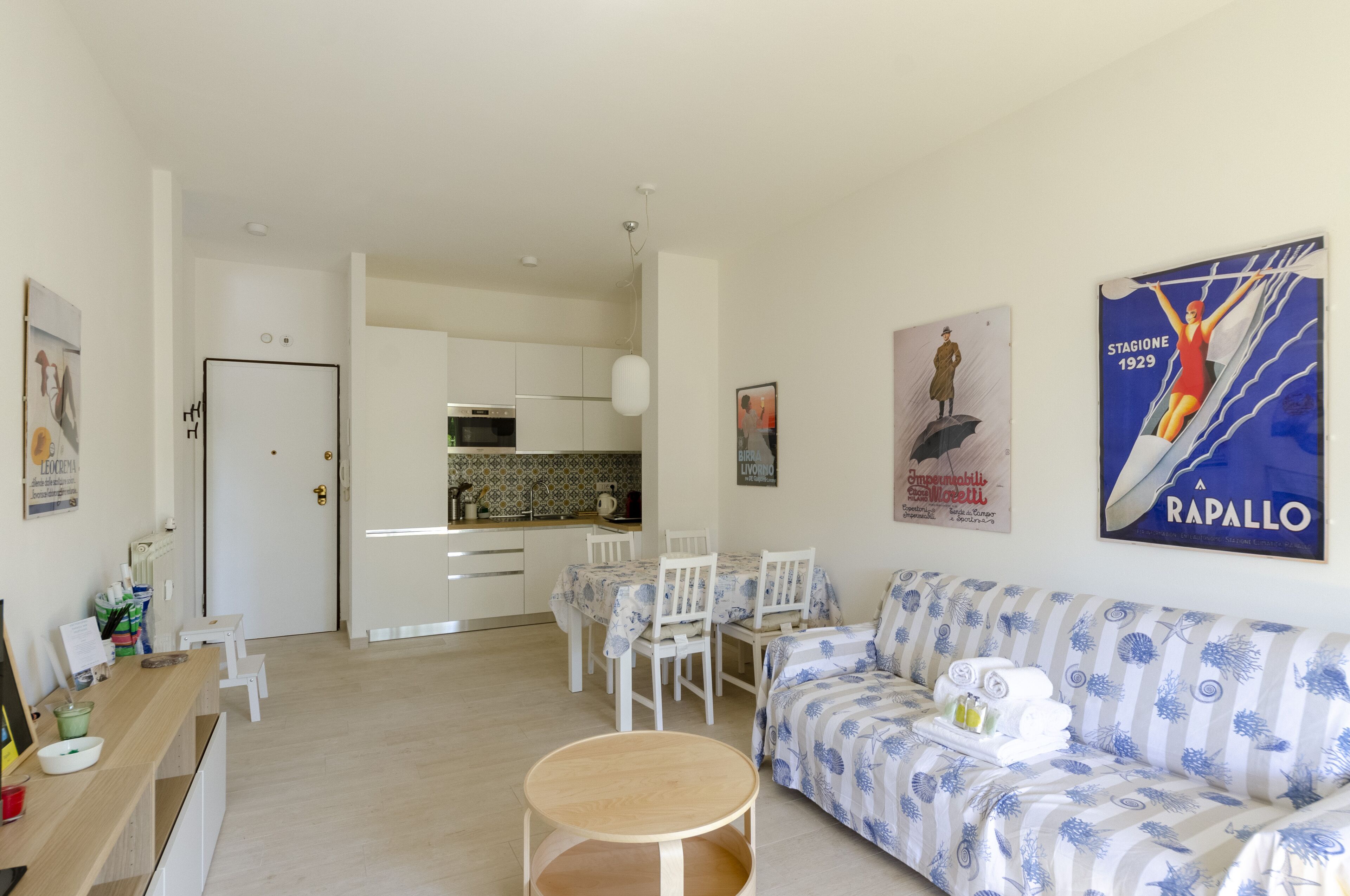 Una sala moderna con un sofá, una mesa de comedor y sillas. Se observa una zona de cocina al fondo con armarios blancos y un salpicadero de azulejos.