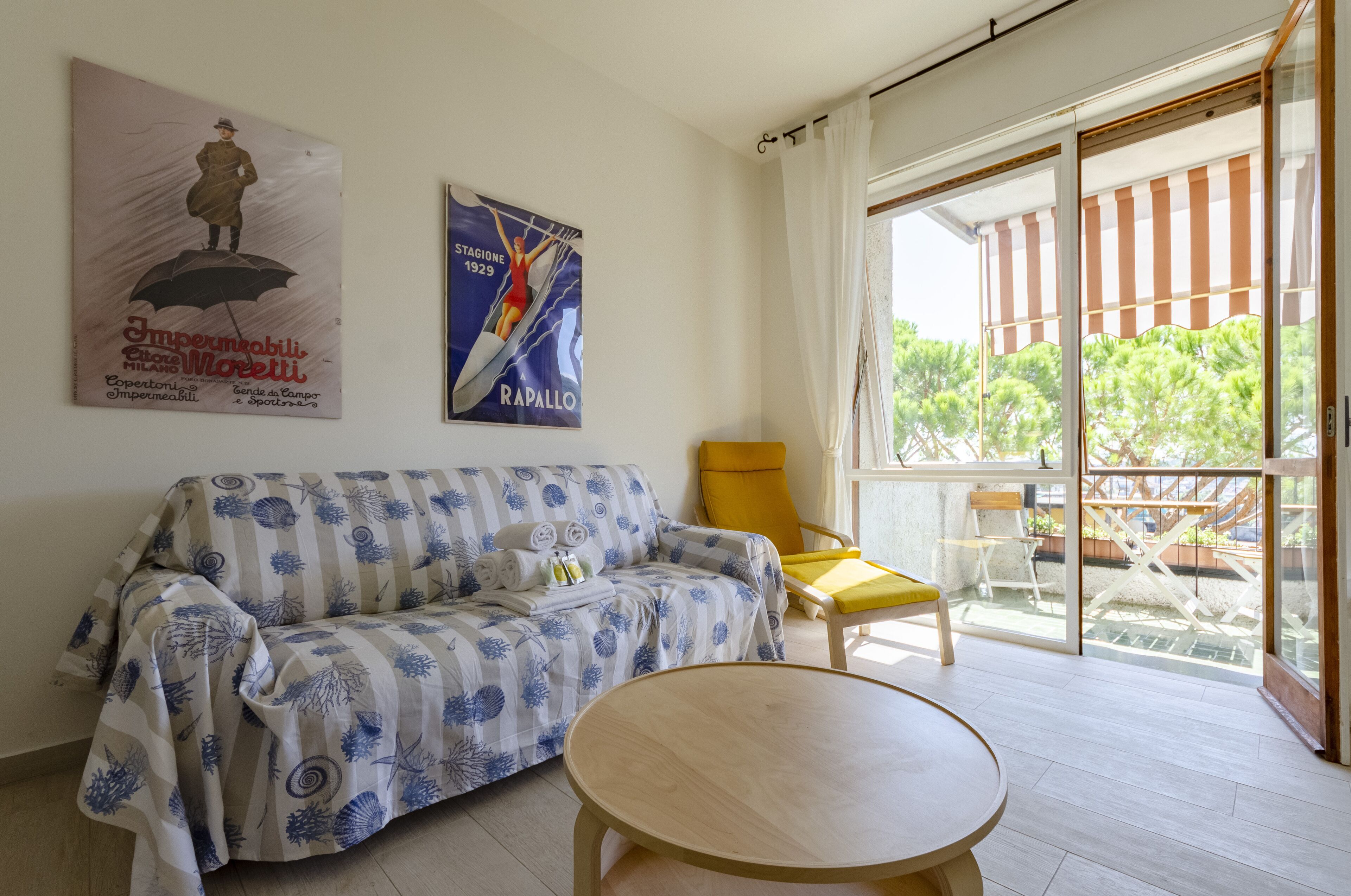 A living room with a sofa, a yellow armchair, and a round table. There are two posters on the wall and a sliding glass door leading to an outdoor area.