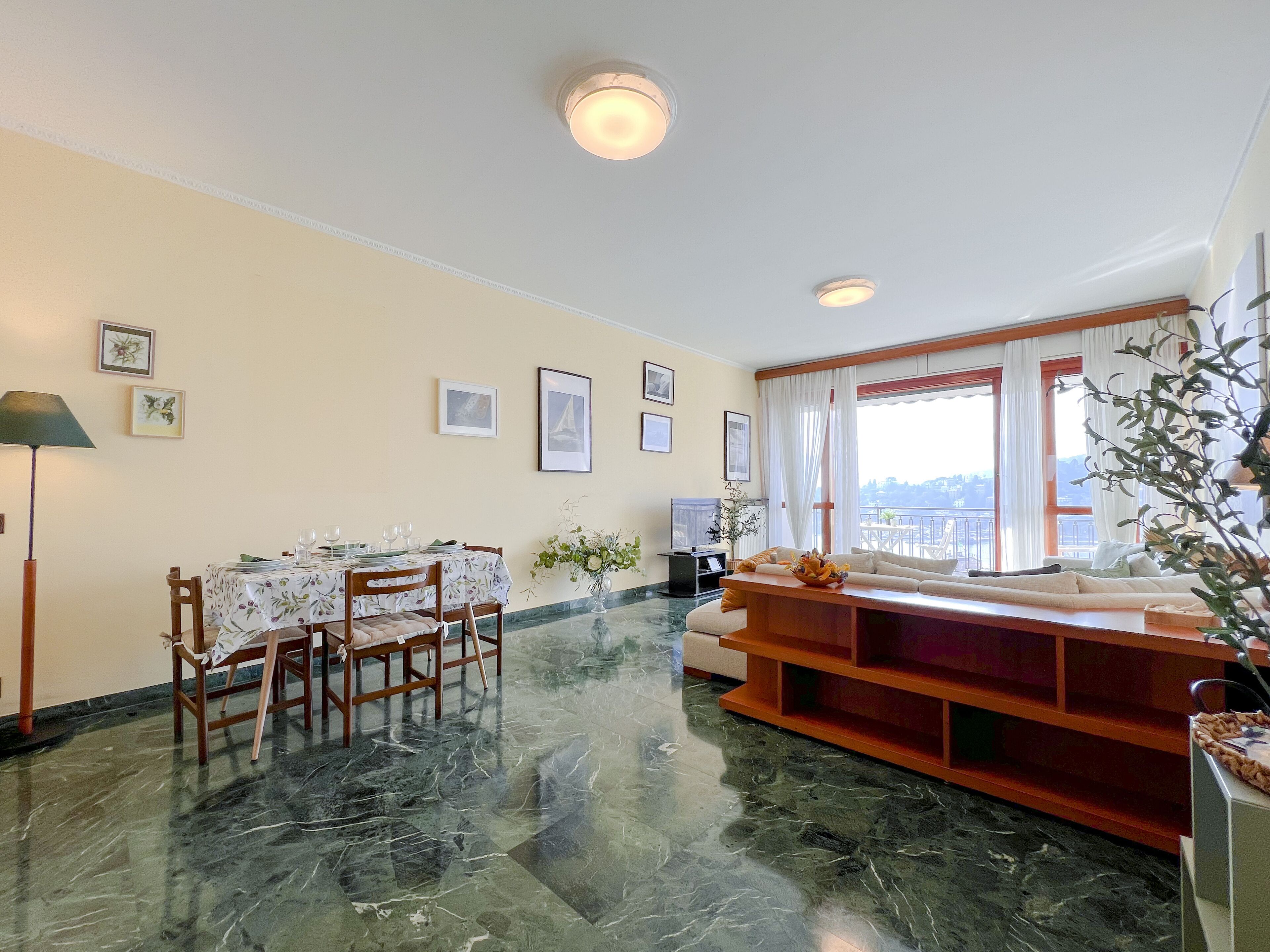 A living room with a dining area, marble flooring, a sofa, a TV, and a balcony with a view.